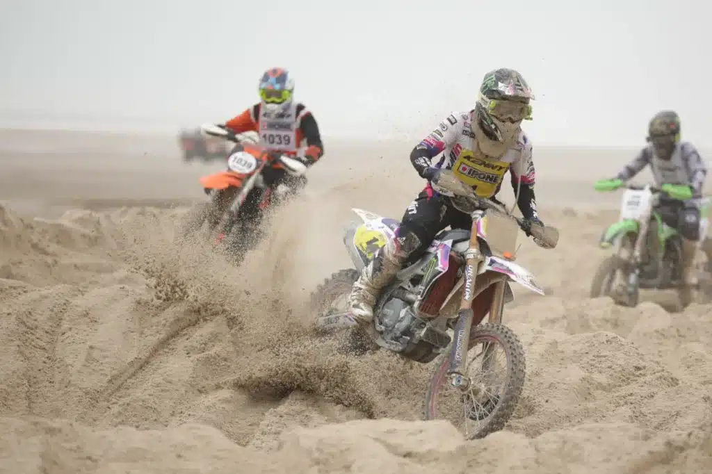 Motocross en action, projetant du sable lors d'une course, avec un gros ballon en arrière-plan affichant le logo de '3AS RACING', le slogan 'L'expert pièces & accessoires moto / quad' visible en-dessous.