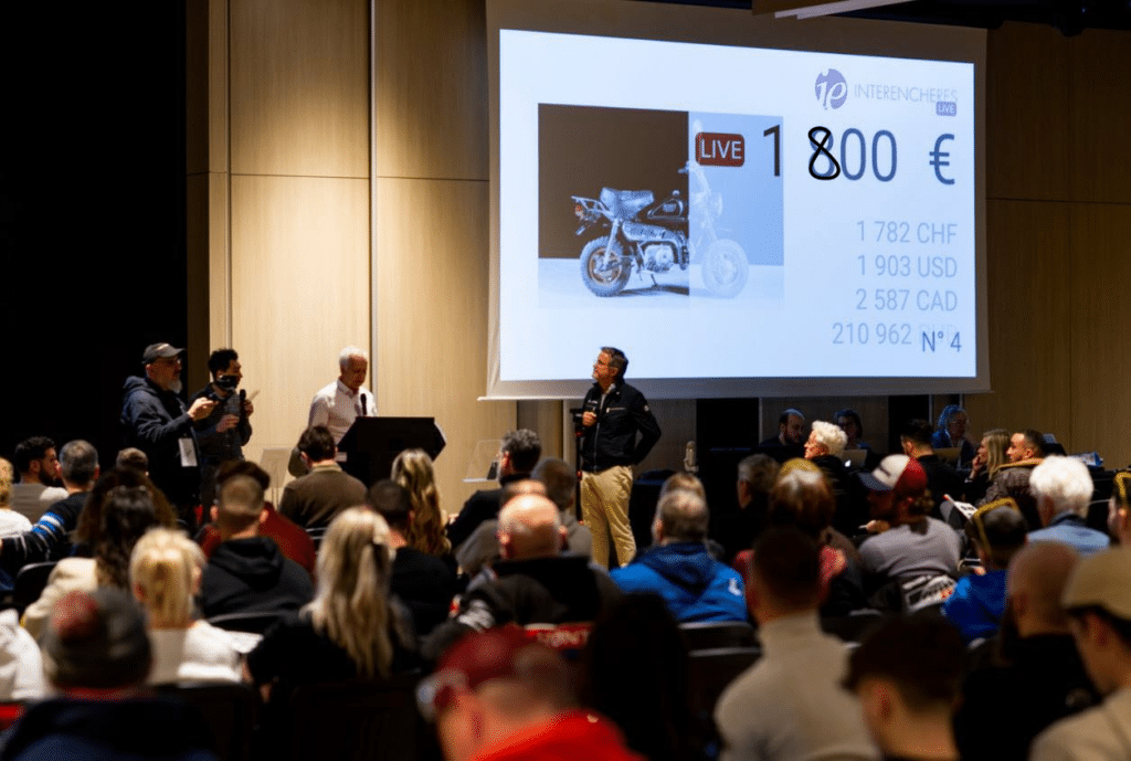 Salle de vente aux enchères avec des participants observant une offre en direct pour une moto, affichée à l'écran avec des prix en EUR, CHF, USD et CAD.