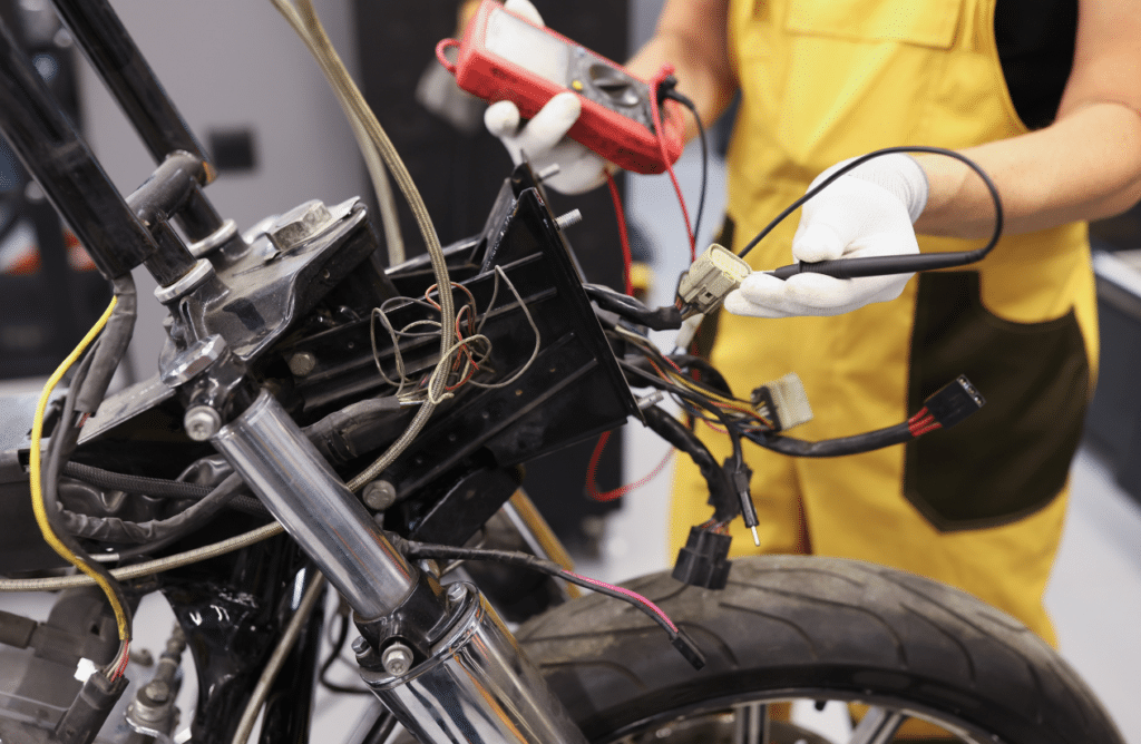 L'image montre un technicien en combinaison de travail jaune, effectuant une maintenance électrique sur une moto, en utilisant un multimètre pour tester la tension d'un composant. Les mains gantées manipulent soigneusement les câbles et connecteurs