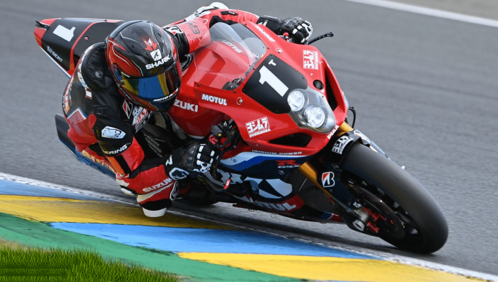 Un pilote de course sur une moto Suzuki numéro 1, vêtu d'une combinaison rouge et noire, en pleine action sur un circuit de course, inclinant fortement dans un virage marqué par des bordures bleues et jaunes.