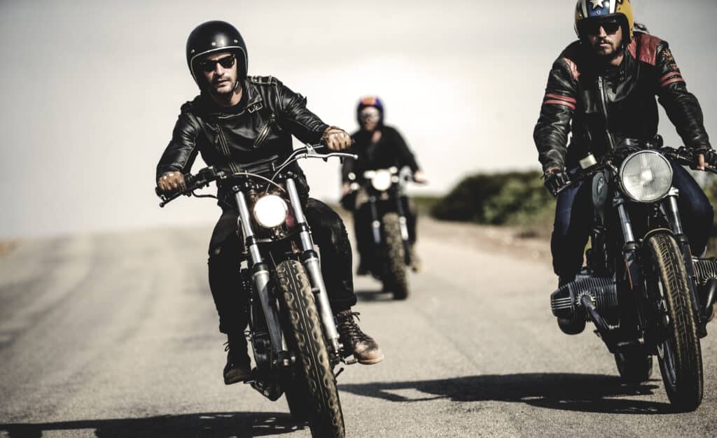 Three men wearing open face crash helmets and sunglasses riding cafe racer motorcycles along rural road.