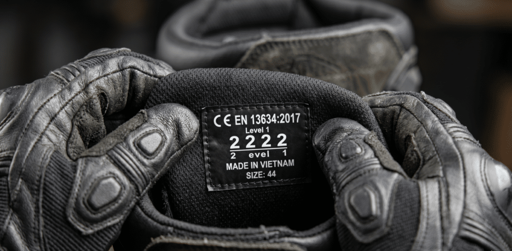 close-up macro shot of a motorcycle boot inner label showing CE EN 13634 certification numbers "2222" in white text on black background, held between two gloved hands, shallow depth of field, sharp focus on label, studio lighting, photorealistic, no watermark