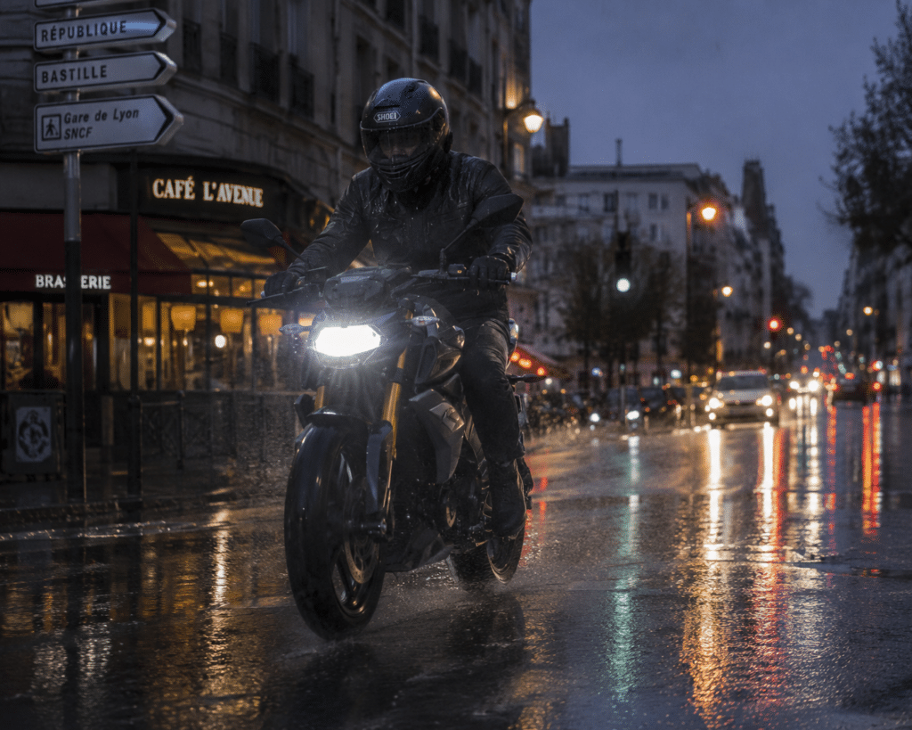 photorealistic, motorcyclist riding through a rain-soaked French urban street at twilight, headlight reflecting on wet asphalt