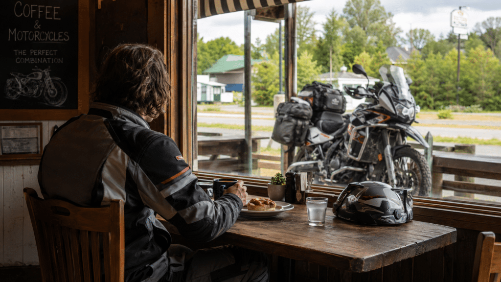 photorealistic, adventure motorcyclist on a rest break eating and drinking coffee at a roadside cafe, helmet on table, gear on, loaded adventure motorcycle visible through window, sense of responsible fatigue management before continuing journey, no face visible, no watermark