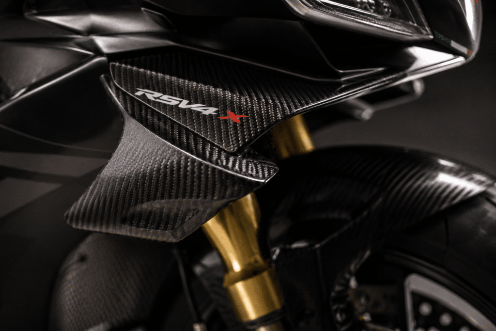 photorealistic extreme close-up of Aprilia Tuono V4 X front aerodynamic carbon winglet, dramatic studio lighting revealing carbon weave texture and engineering detail, sense of MotoGP technology transfer to production track bike, sharp technical focus, no watermark